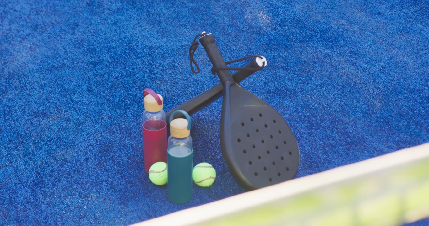 Kamera fährt heran und enthüllt Schläger, Flaschen und Bälle auf blauem Spielfeld, um Details zu zeigen.