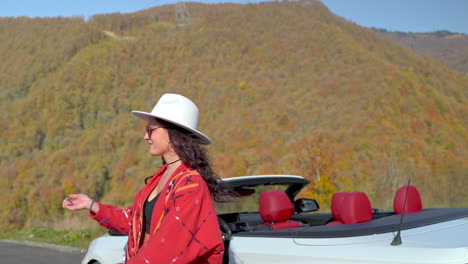 woman in a red jacket and white hat by a convertible car in autumn mountains