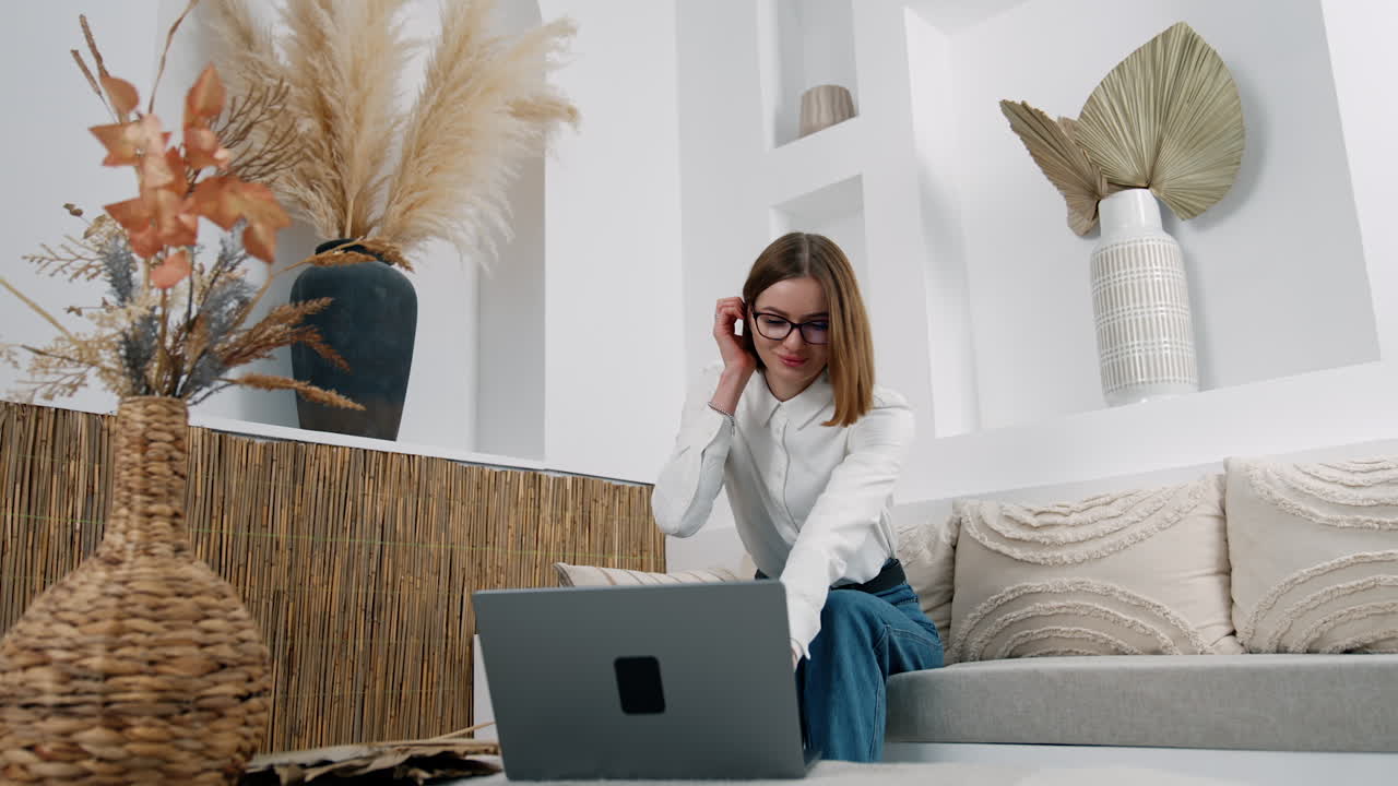 Brunette girl leaning to her laptop standing on the coffee table. Lady looks at the screen smiling slightly to camera. Low angle view.