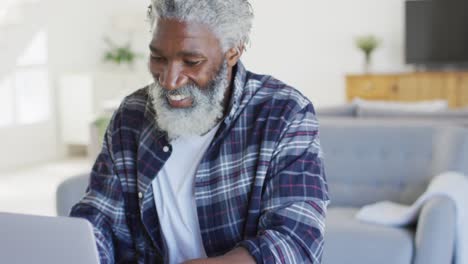 african american senior man sitting by a table, using laptop and smiling, social distancing and self