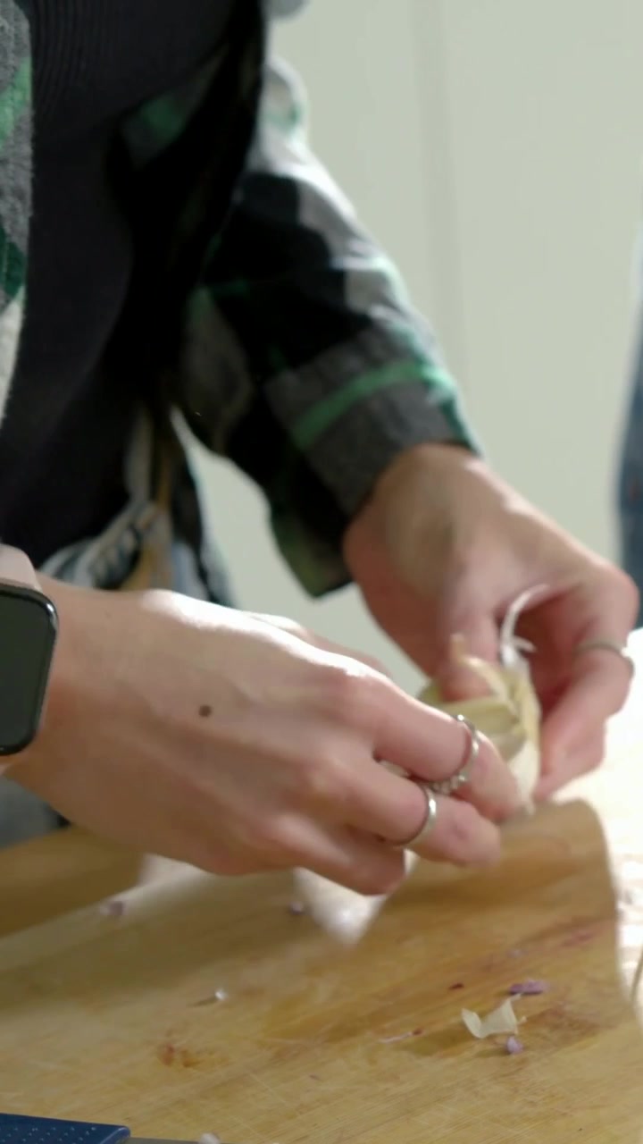 Video verticale: Una donna in plaid afferra un bulbo d'aglio e sbuccia uno spicchio sul bancone, mentre un'amica affetta per cucinare.