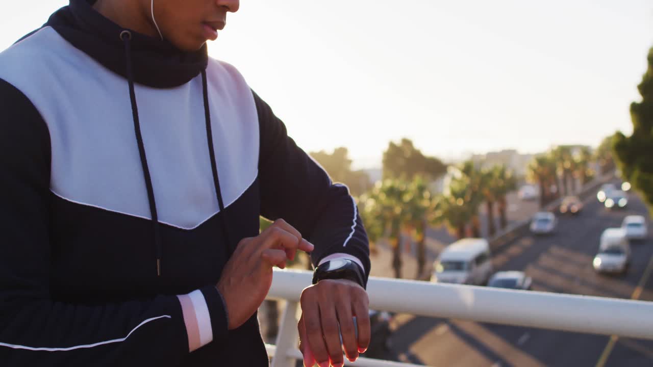 Midsection of african american man wearing earphones exercising on footbridge checking smartwatch