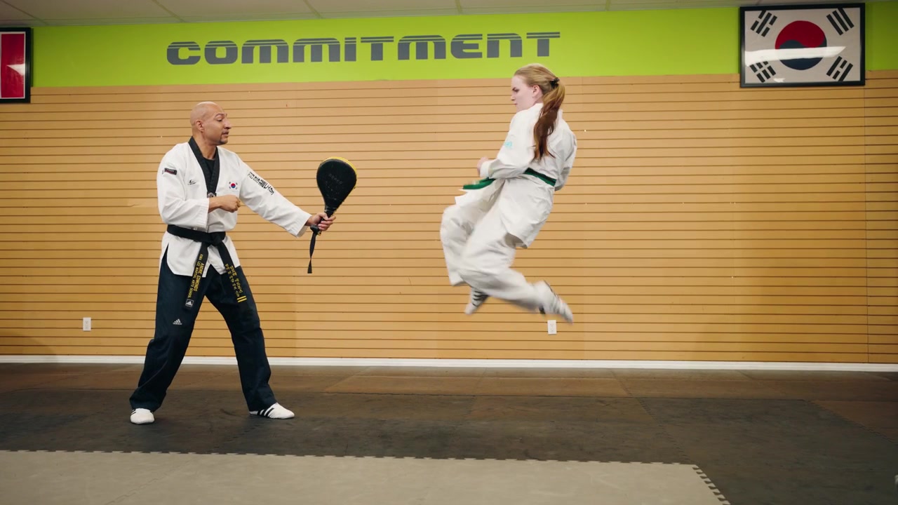 En ung kvindelig kampsportsudøver inden for Taekwondo og Karate udfører to hoppende slag og træner et roterende dobbelt højt spark mod en pude med en sortbælte-mester i et trænings- og styrkecenter.