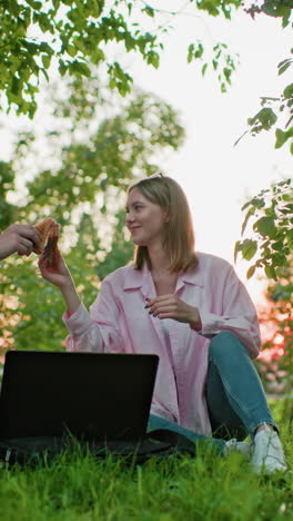 brother and sister seated outdoors with laptop in front making a toast with bread in hand, enjoying meal together in natural sunlight filtering through trees
