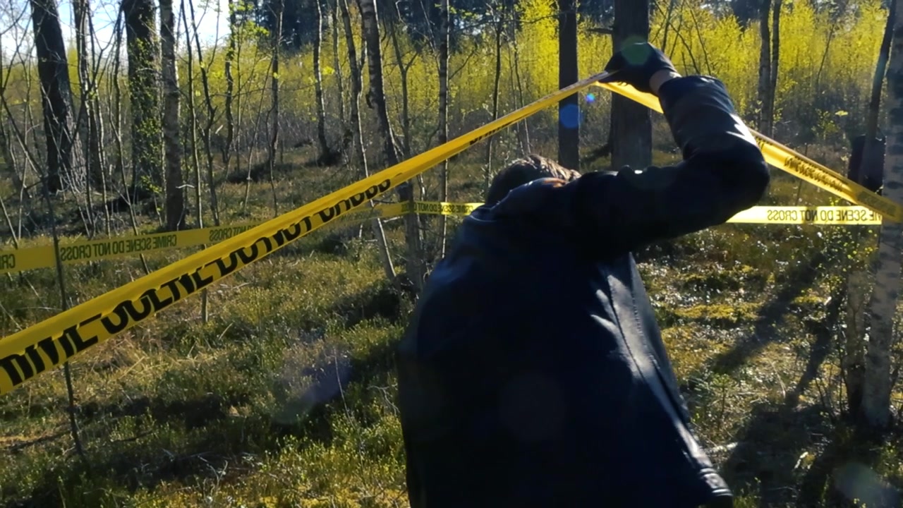 Male police detective wearing a black leather jacket and gloves crossing a yellow tape to enter an outdoor crime scene in a sunlit forest, carefully inspecting the area for evidence