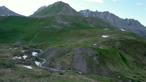 Imágenes-Aéreas-De-Drones-Acercándose-Lentamente-A-Una-Cabaña-Alpina-Suiza-Ubicada-Entre-Las-Laderas-Cubiertas-De-Hierba-En-Un-Paisaje-Montañoso-Con-Parches-Residuales-De-Nieve-Y-Prados-Alpinos-En-Suiza