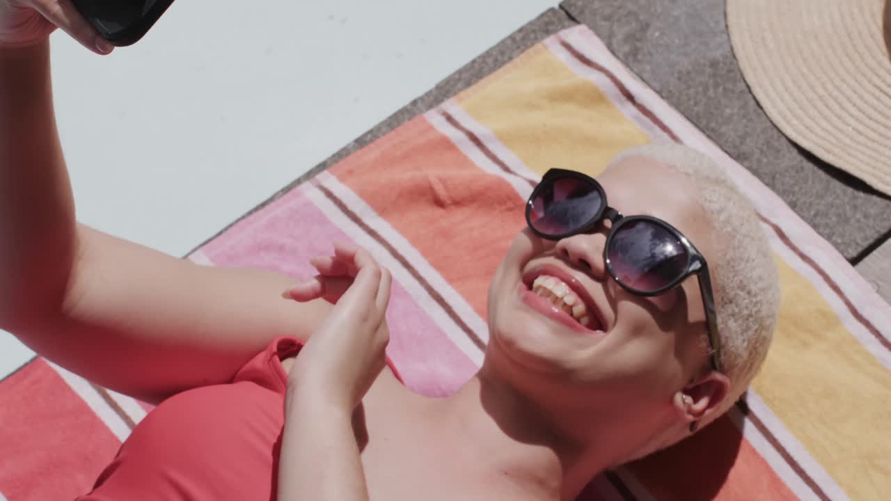 Happy biracial woman with sunglasses using smartphone and sunbathing on towel in slow motion
