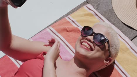 happy biracial woman with sunglasses using smartphone and sunbathing on towel in slow motion