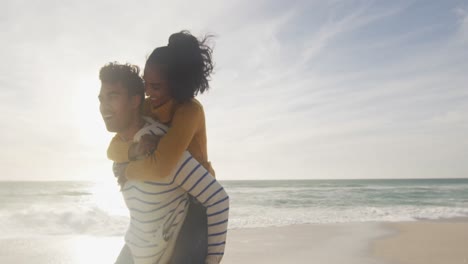 hombre hispano feliz llevando a una mujer a hombros y divirtiéndose en la playa