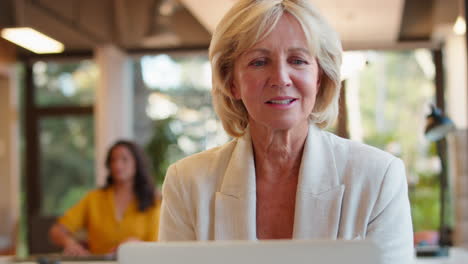 mature businesswoman working on laptop at desk in office pausing to look out of window