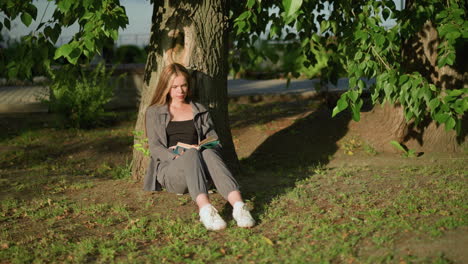 lady sitting outdoors, leaning against tree on grassy field, reading book under warm sunlight, tree leaves sway gently in breeze, shadows cast on ground, background features greenery