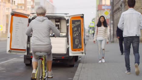 blonde frau fährt fahrrad, hält vor einem cafe-truck an und bittet den kellner um einen kaffee