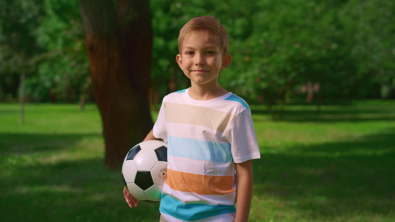 Criança pequena com bola sorrindo na natureza. Retrato de jovem esportista fofo no parque.