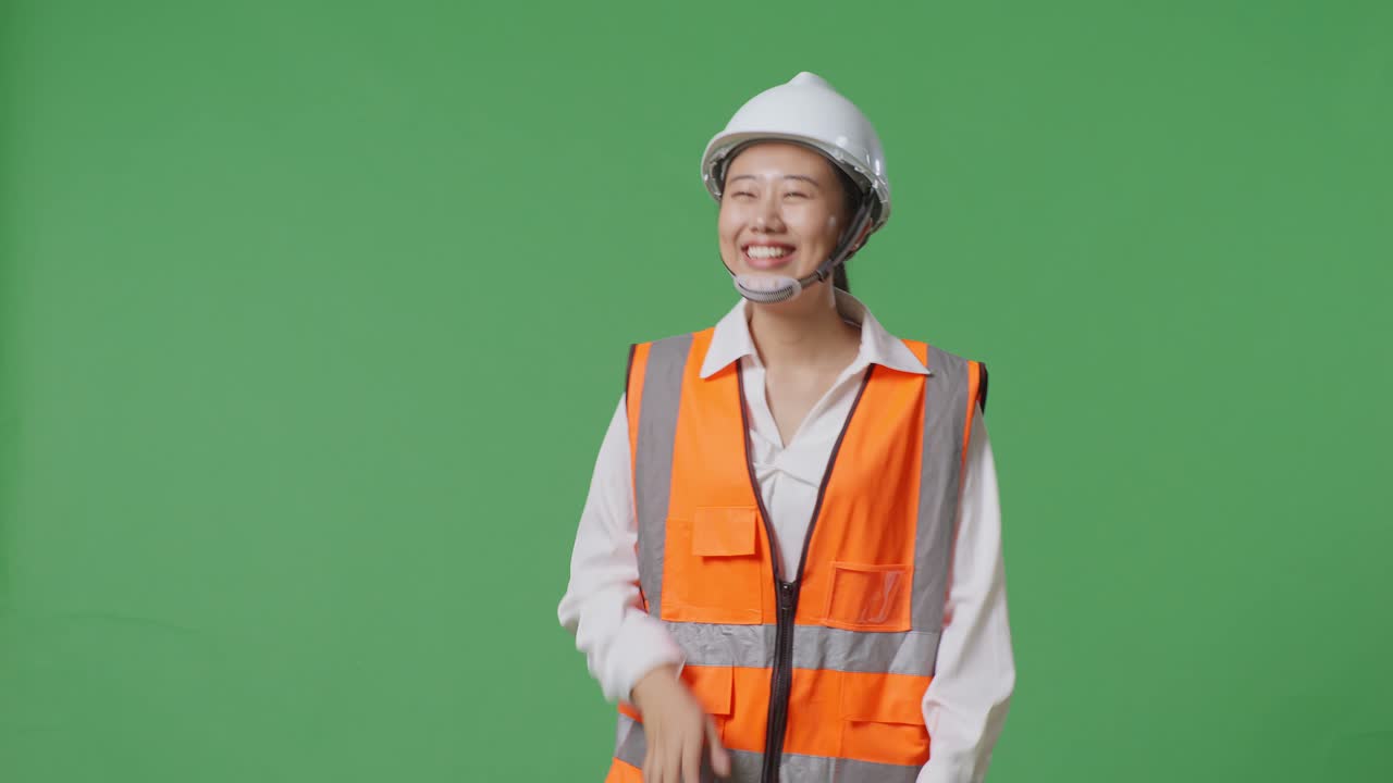 안전 헬을 입은 아시아 여성 엔지니어가 녹색 스크린 배경 스튜디오에서 는 동안 손을 흔들고 있습니다.
