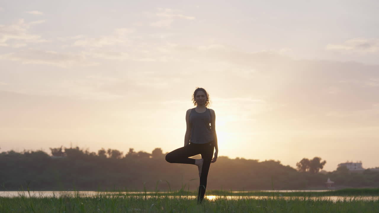 Un grandangolo di una donna sud-asiatica che esegue la posizione dell'albero in equilibrio su una sponda erbosa del lago all'alba, in controluce rispetto al cielo dorato