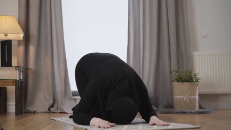 young muslim lady in black traditional hijab bowing down and raising up with hands raised. modern eastern woman praying at home. eastern culture, religion, lifestyle.