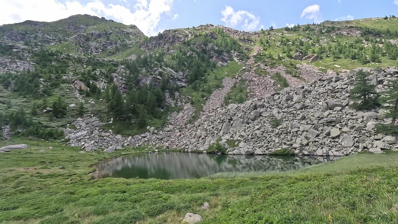 Still alpine lake nestled beneath rocky slopes and conifer-covered ridges in Alpe Veglia, Italy, under a bright summer sky