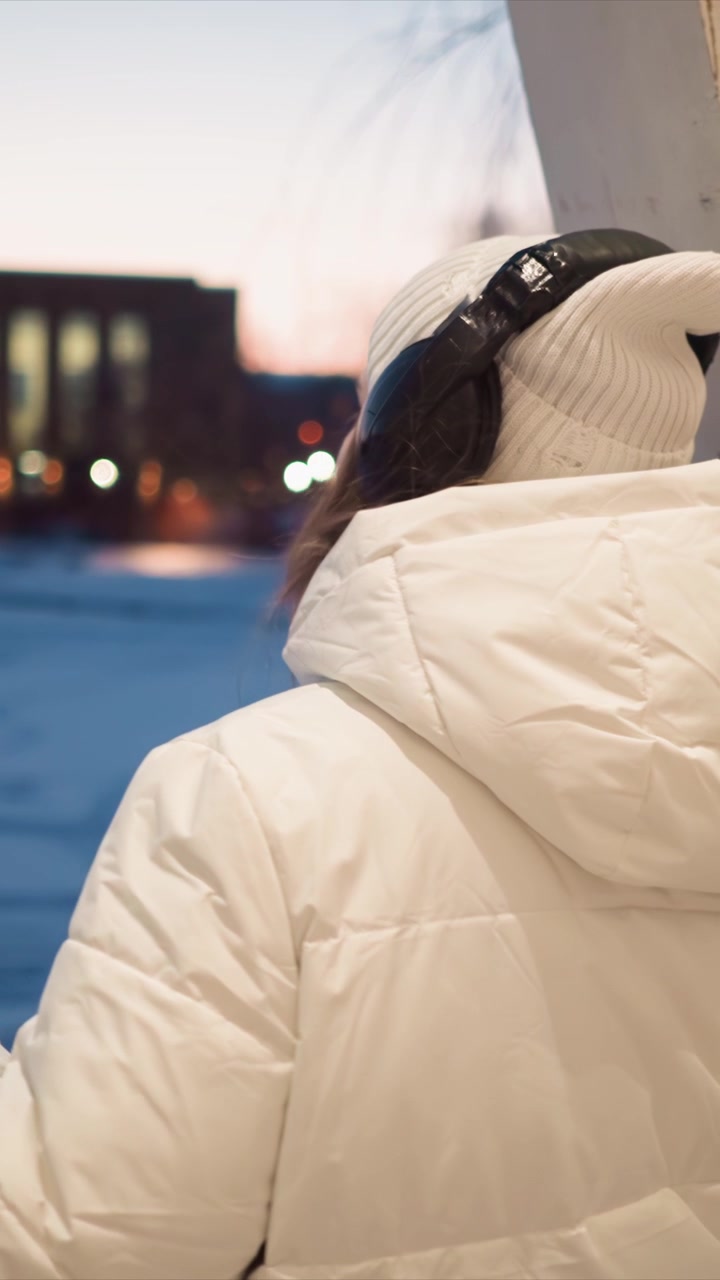 Woman in white winter coat and knit hat stands outdoors facing away from camera. background shows urban setting with blurred lights and buildings during twilight or early evening. Soft