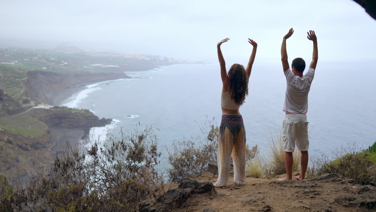 On a cliff's edge, a man and woman raise their hands and breathe in the ocean air during a yoga session, absorbing the serenity