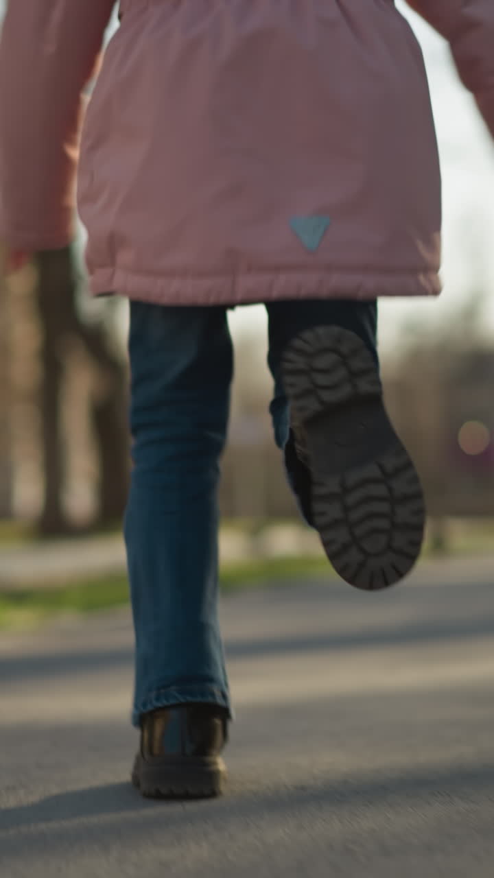 primer plano de las piernas de una joven en movimiento, con pantalones vaqueros azules y zapatos negros, corriendo por un camino de parque iluminado por el sol con árboles borrosos y un fondo de edificio