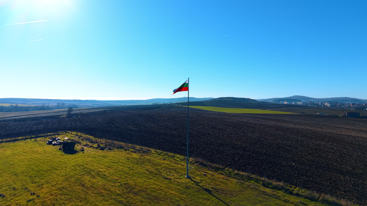 National flag of Bulgaria waving in rural landscape. Bulgarian flag on a high flagpole against blue sky and agricultural fields with sun flare