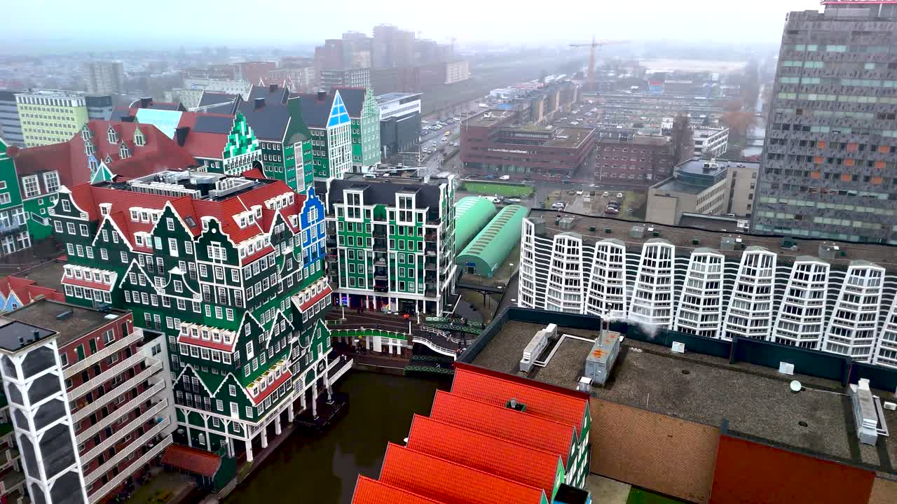 Aerial view shows unique, colorful buildings lining a canal. The area features a mix of modern and traditional architecture, with an overcast sky above