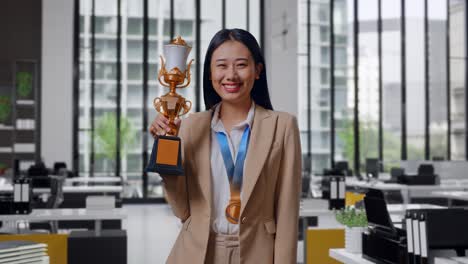 una mujer de negocios asiática en traje lleva una medalla de oro y sostiene un trofeo de oro en su mano como la primera ganadora sonriendo a la cámara en la oficina