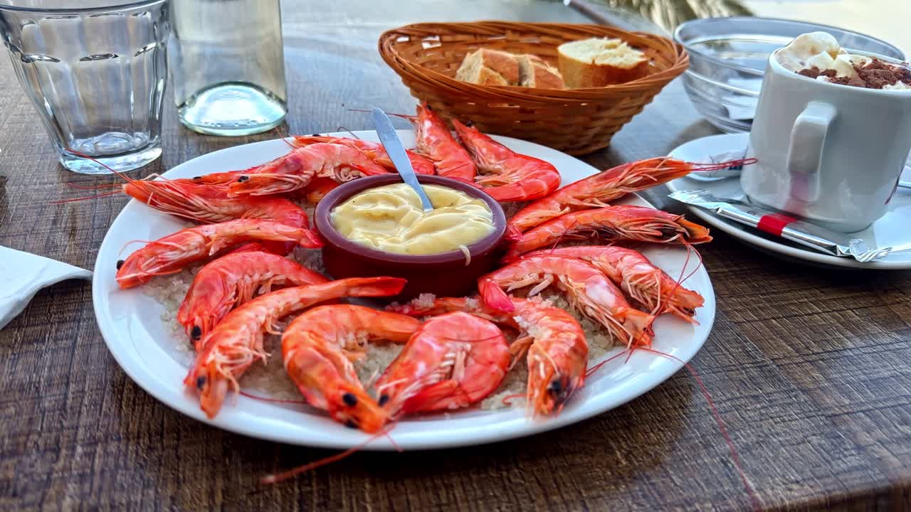 Plate of cooked shrimp served on ice with dipping sauce at an outdoor restaurant table, creating a fresh seafood dining scene with bread and drinks