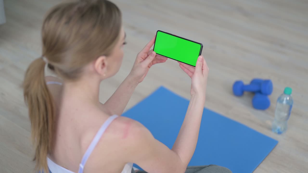 요가 매트 에 크로마 키 스크린 을 가진 스마트폰 을 보고 있는 젊은 여성 의 뒷면