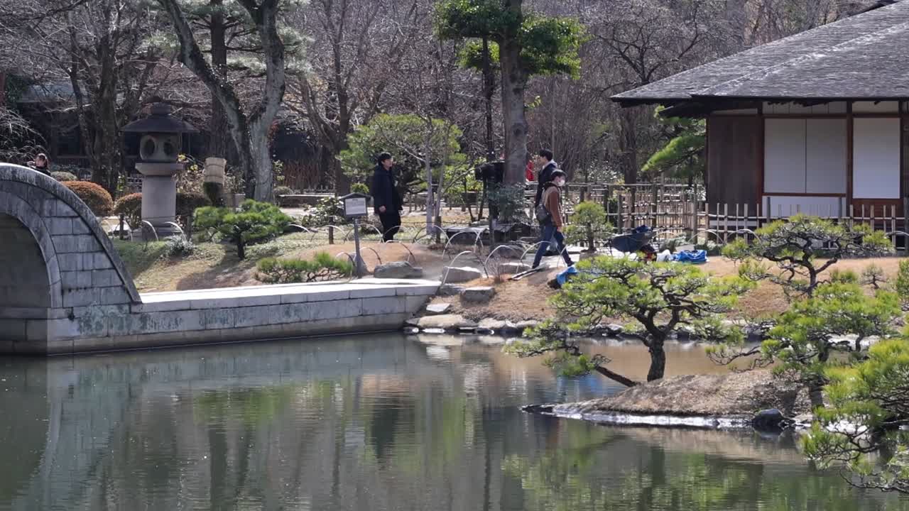 A peaceful garden scene featuring a stone path, traditional building, and reflective pond under a clear sky.