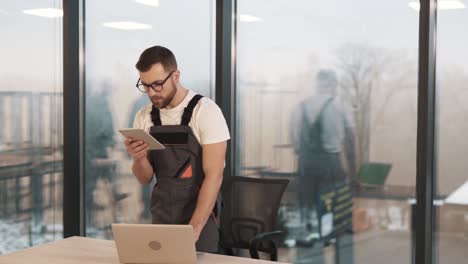 un trabajador uniformado con una computadora portátil y una tableta en una nueva oficina moderna está instalando software, alarma, videovigilancia y equipo de acceso seguro en el contexto de ventanas panorámicas