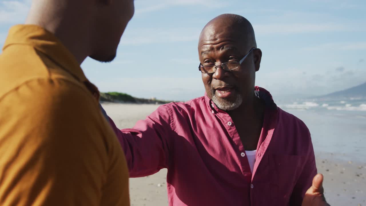padre afroamericano sentado en la playa con su hijo adolescente, poniendo la mano en su hombro y hablando