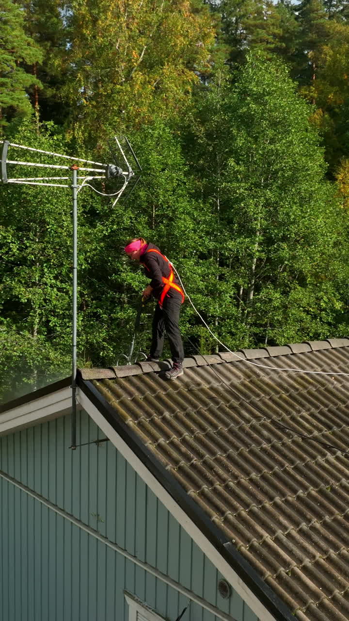 Aerial portrait rotating around a woman pressure washing a house roof, sunny day