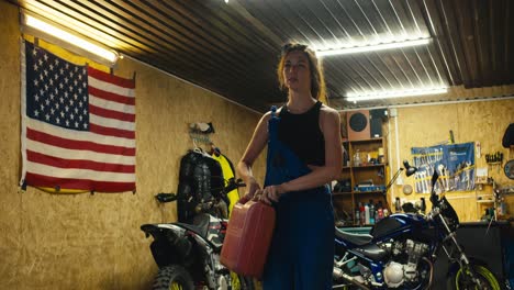 a tired girl mechanic in a black top and blue overalls comes out of her garage workshop along with a canister of water and pours herself near the entrance to the garage. an impromptu shower after a hard day in the workshop