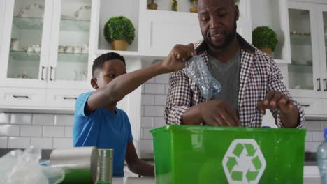 happy african american father and son standing in kitchen putting plastic rubbish in recycling box