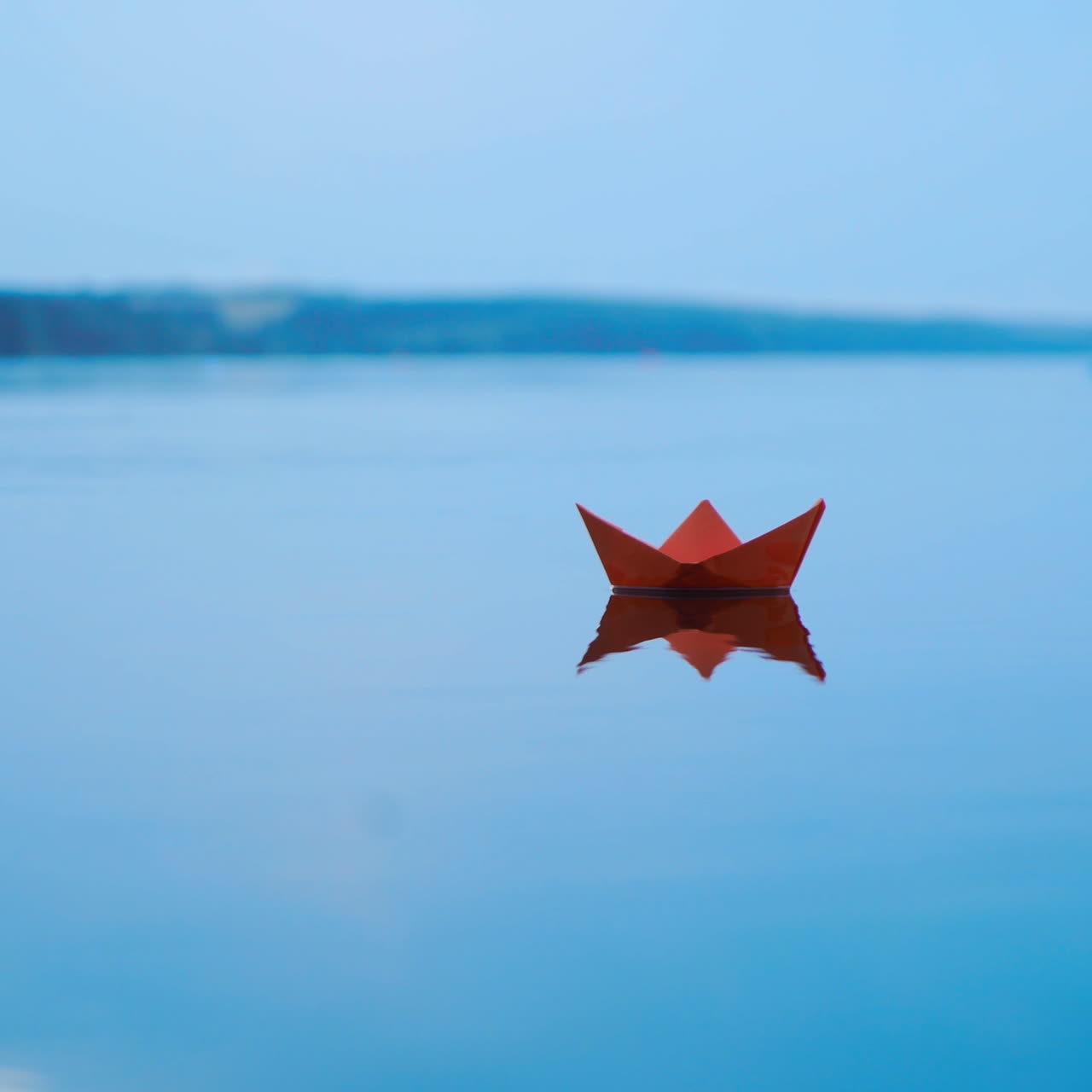 Red paper boat sailing alone on blue water surface under the blue sky. Origami ship on the river at sunset.
