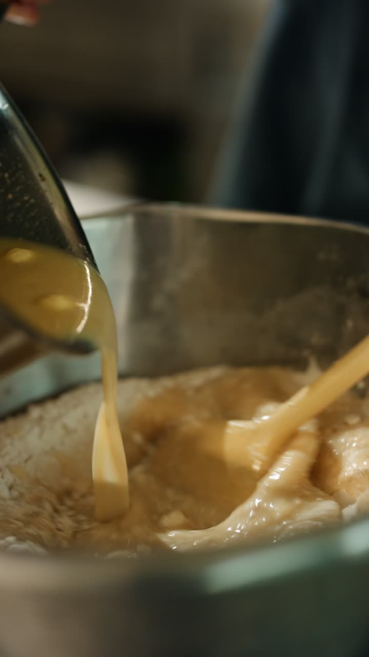Preparing Buchteln yeast rolls, pouring liquid ingredients into a metal bowl with flour and a wooden spoon, making Austrian sweet pastry dough in the kitchen, vertical shot, real time