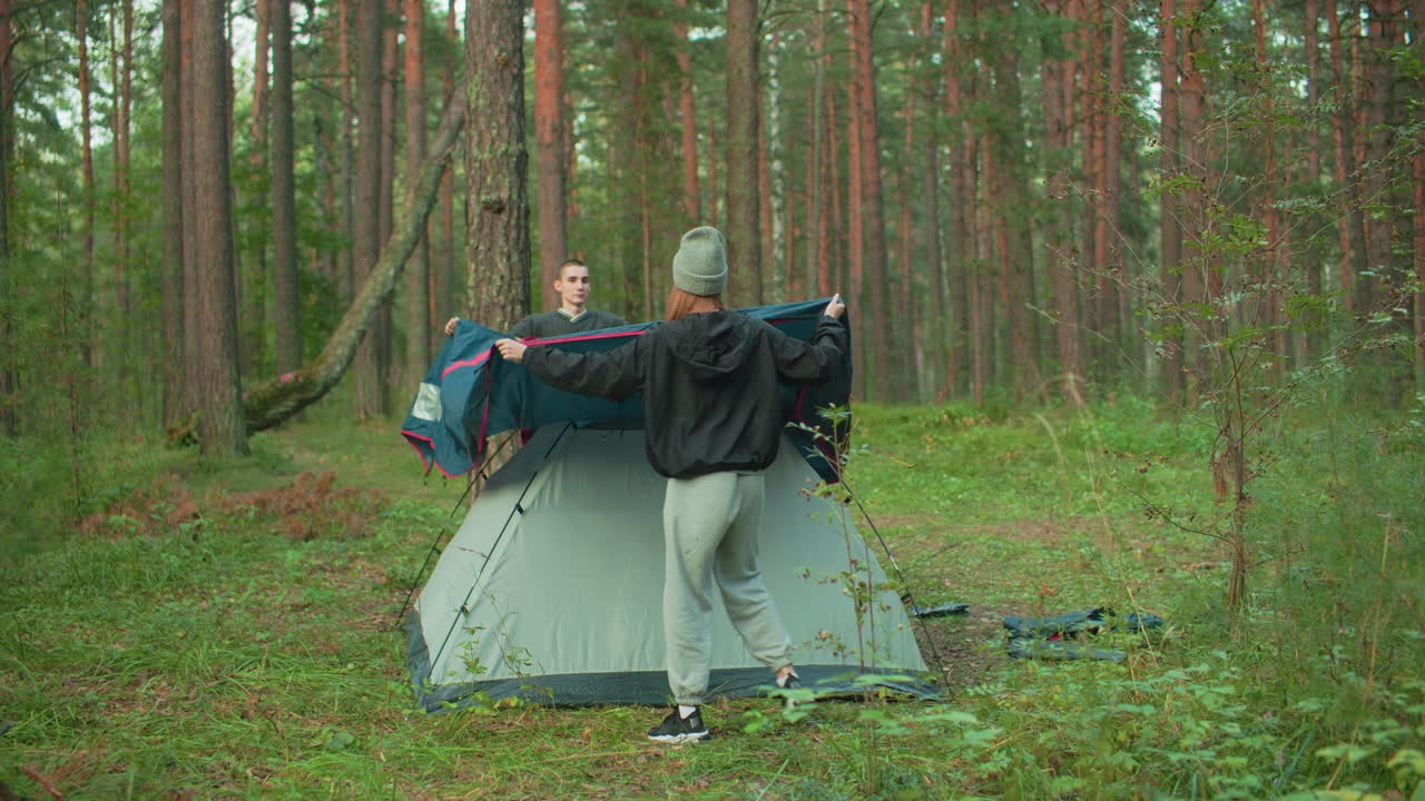 Young camper carefully places tent cover over pitched tent surrounded by dense forest trees and green undergrowth, preparing shelter in peaceful outdoor environment during nature retreat