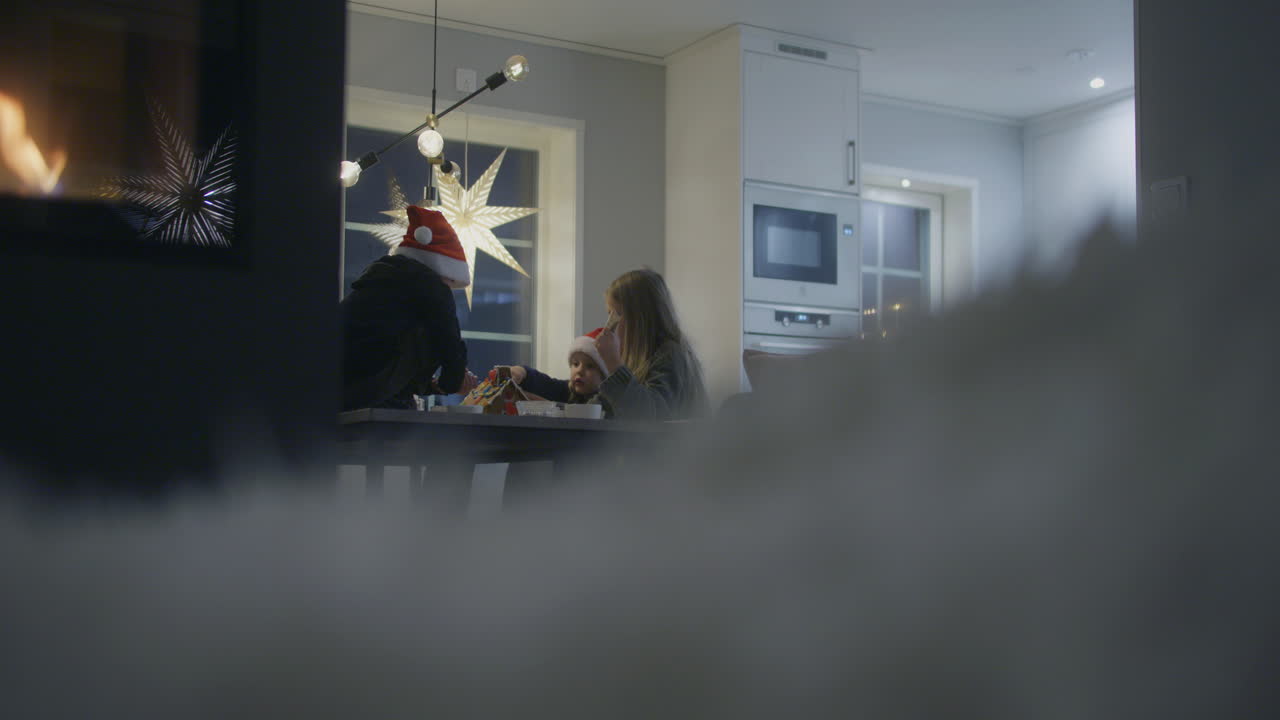 siblings build a gingerbread house at the kitchen table enjoying the fireplace nearby