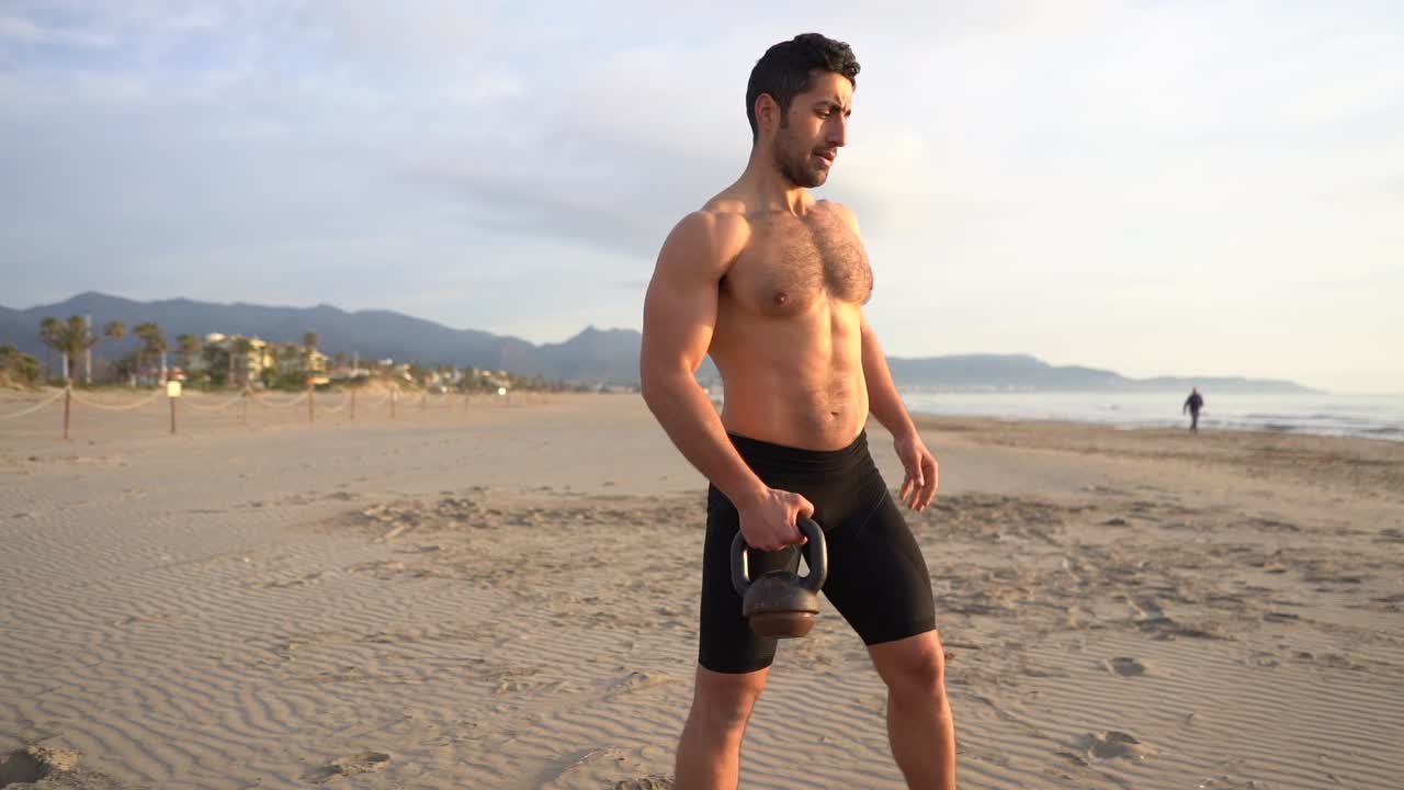 hombre musculoso entrenando con pesas rusas y sin camisa en la playa al amanecer
