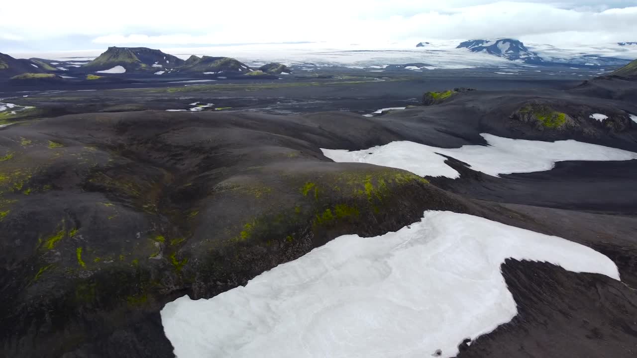 Aerial drone slowly flying forward over Iceland volcanic dark brown and mossy white snow covered landscape terrain with large mountains visible in the background. Clouds are around the steep mountains