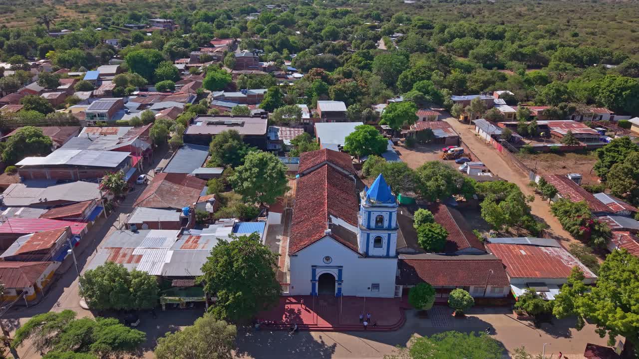 Drone voa para trás a partir da icônica Capilla Santa Bárbara azul e branca em Villavieja, Colômbia. A praça da cidade está escondida por exuberantes árvores verdes, destacando esta charmosa entrada para o Deserto de Tatacoa.