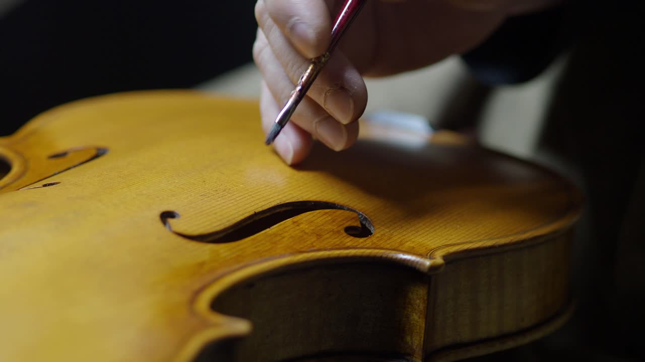 Close-up of a luthier using a fine-tipped burnisher to perfect the f-hole on a spruce violin top. Hand-crafted maple body, visible grain, and varnish application showcase precision craftsmanship