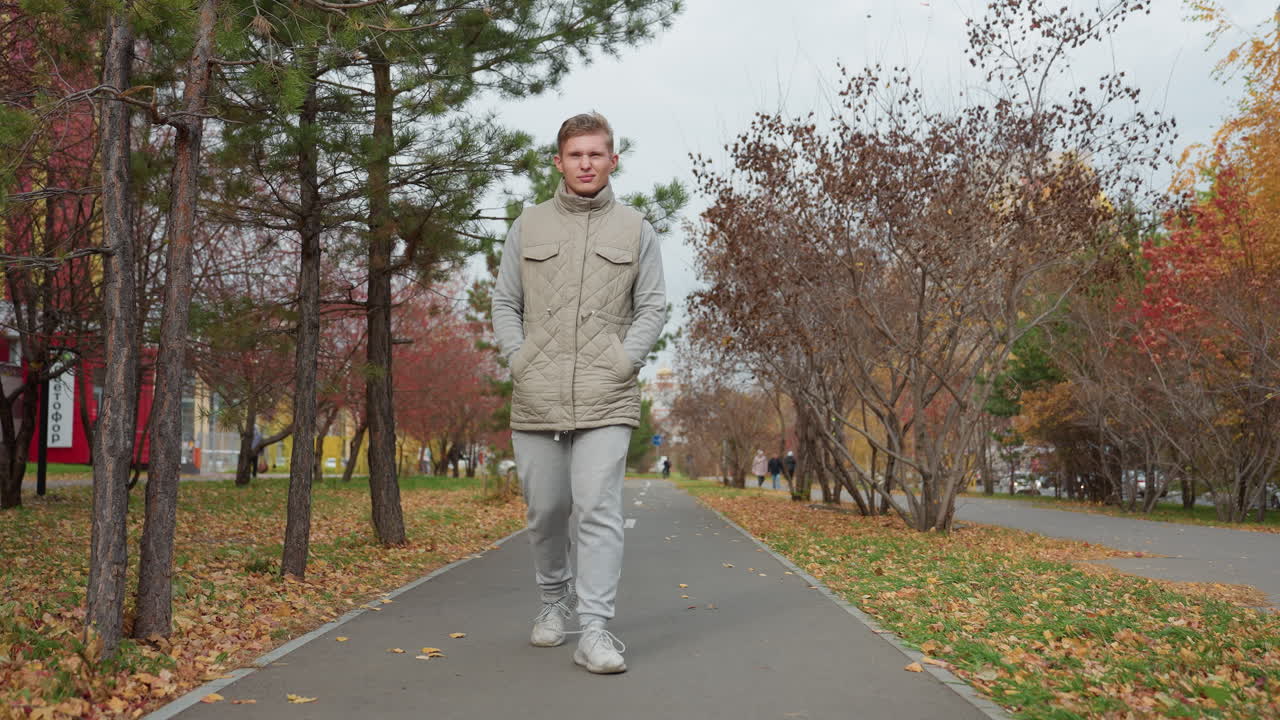 Young man walking alone on paved path in autumn park surrounded by colorful trees with fallen leaves covering ground as light wind blows foliage, cars moving in background on cloudy day