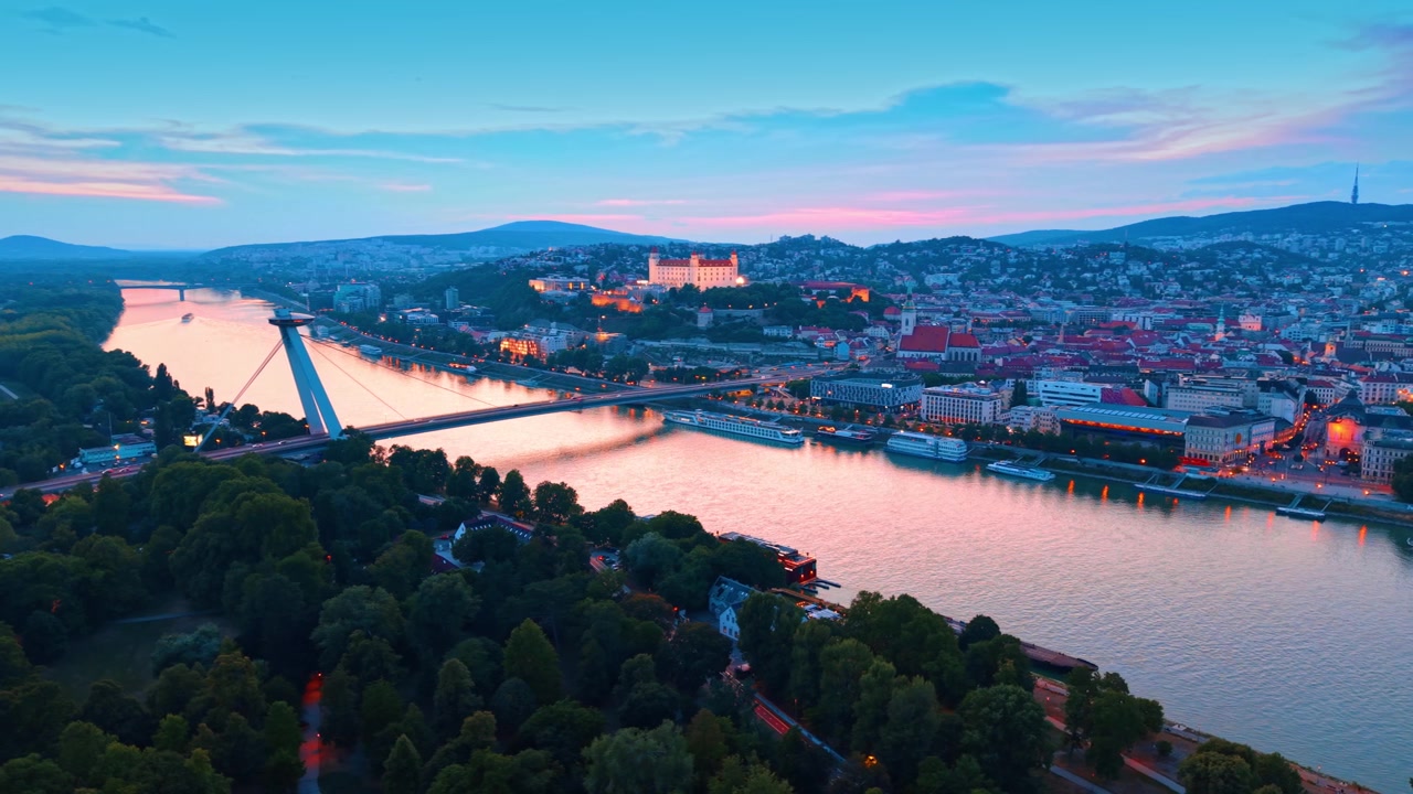 Veduta aerea panoramica di un parco verde, fiume, ponte e castello illuminato sotto il cielo del tramonto nella città di Bratislava