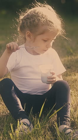 little blond lady in white t-shirt black leggings eats yogurt with spoon sitting on meadow in trees shadow slow motion