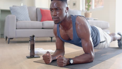 tired african american man doing plank, using headphones and smartphone in living room, slow motion