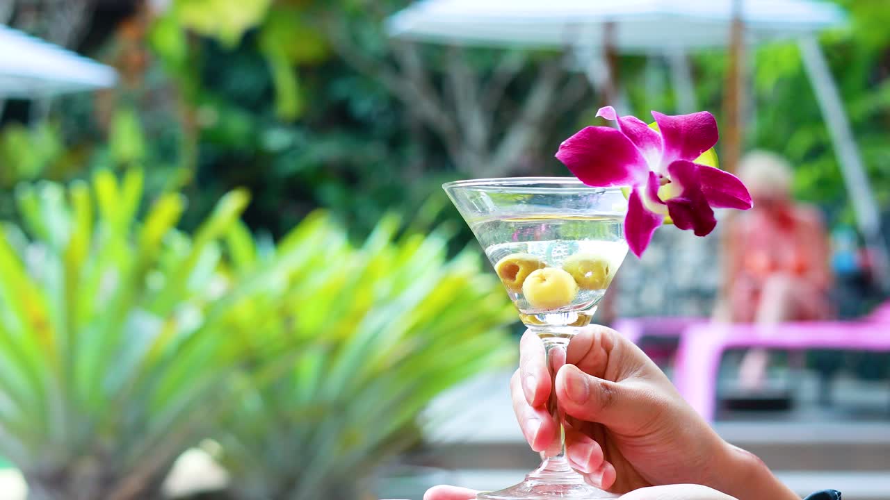 손은 올리브와 꽃으로 마르티니 글래스를 들고, 활기찬 수영장 배경에 설정