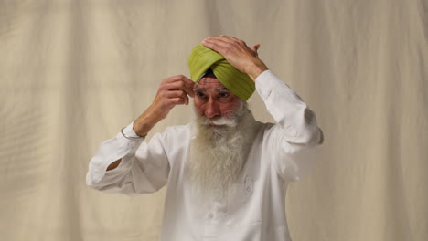 Fotografía-De-Estudio-De-Un-Hombre-Sikh-Mayor-Con-Barba-Que-Usa-Una-Aguja-De-Salai-Al-Ponerse-Un-Turbante-Contra-Un-Fondo-Liso