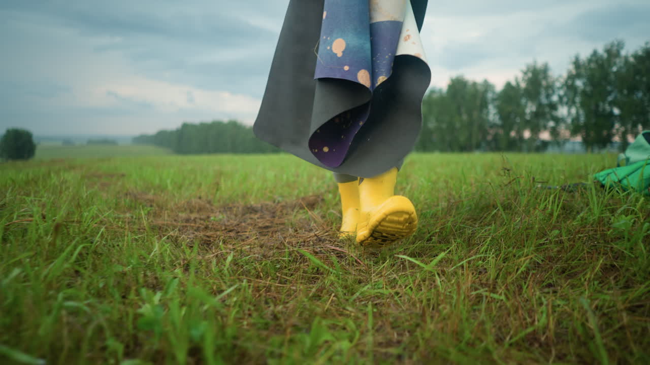 노란색 부츠를 입고 다리를 들고 다채로운 매트를 들고 잔디에서 는 개인의 다리 시각, 푸른 레인코트와 가방은 광활한 의 바닥에서 뒤에 보입니다.